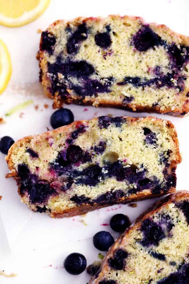 Blueberry Zucchini Bread with a Lemon Glaze will be one of the best quick breads you EVER make! Perfectly moist with two cups of zucchini hidden inside and bursting with fresh blueberries. The lemon glaze it the perfect finishing touch! Blueberry Zucchini Bread with a Lemon Glaze that has been sliced and fresh blueberries next to it.