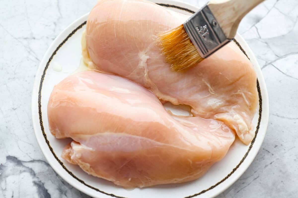 With this Air Fyer Chicken recipe, I am going to show you how to make the most insanely tender and juicy chicken! The secret? The incredible seasonings! They create a nice golden, crispy outside with a juicy, flavor-packed inside! Someone brushing olive oil onto the two raw chicken breasts.